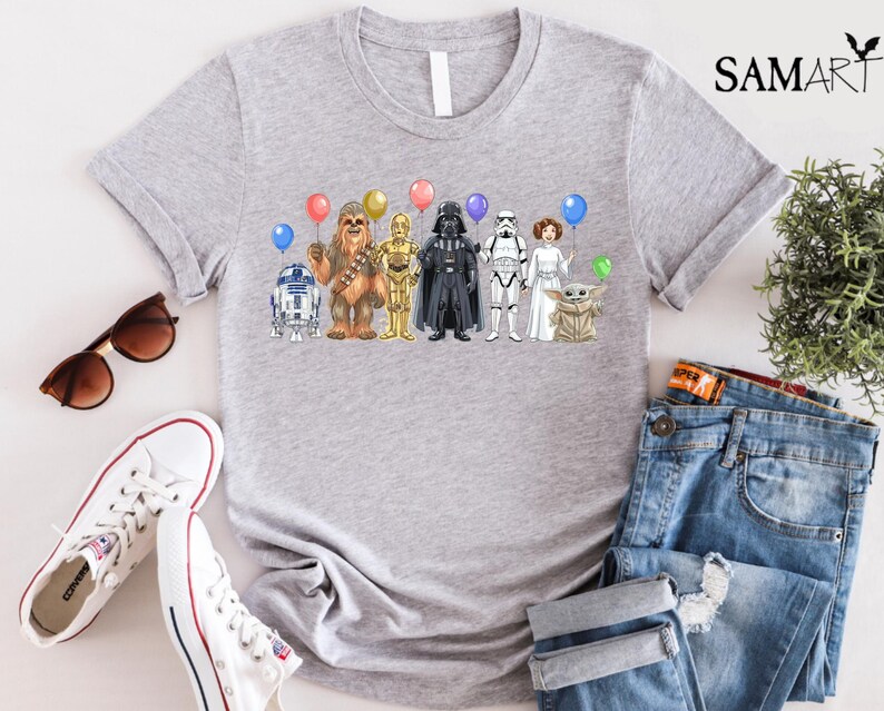 Op de afbeelding: Een lichtgrijs t-shirt met een kleurrijke afbeelding van Star Wars-personages die ballonnen vasthouden. De personages zijn onder meer R2-D2, Chewbacca, C-3PO, Darth Vader, een Stormtrooper, prinses Leia en Grogu. Het shirt wordt gecombineerd met een blauwe jeans en witte sneakers.