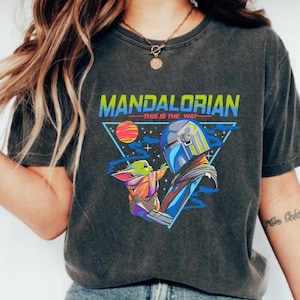 May include: A black t-shirt with a retro-style graphic of a Mandalorian character holding a small green creature. The text "Mandalorian - This is the Way -" is printed on the shirt.