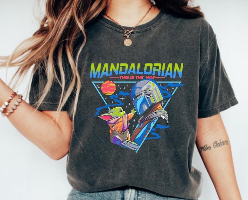 May include: A black t-shirt with a retro-style graphic of a Mandalorian character holding a small green creature. The text "Mandalorian - This is the Way -" is printed on the shirt.