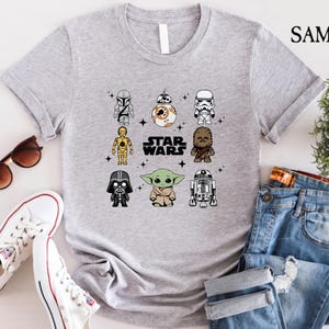 K&ouml;nnte beinhalten: Hellgraues T-Shirt mit einem Star Wars-Motiv, das Cartoon-Charaktere wie Darth Vader und Yoda zeigt. Der Text "STAR WARS" befindet sich in der Mitte. Das Shirt wird mit Sonnenbrille, wei&szlig;en Turnschuhen und blauen Jeans kombiniert.