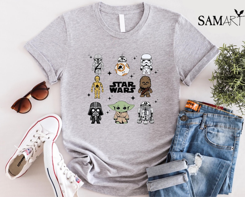 K&ouml;nnte beinhalten: Hellgraues T-Shirt mit einem Star Wars-Motiv, das Cartoon-Charaktere wie Darth Vader und Yoda zeigt. Der Text "STAR WARS" befindet sich in der Mitte. Das Shirt wird mit Sonnenbrille, wei&szlig;en Turnschuhen und blauen Jeans kombiniert.