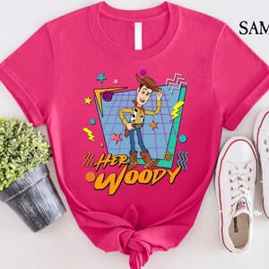 以下が含まれることがあります： トイストーリーのウッディの漫画の画像が描かれた、レトロな90年代のデザインのピンクのTシャツ。Tシャツには「Her Woody」という文字がプリントされています。