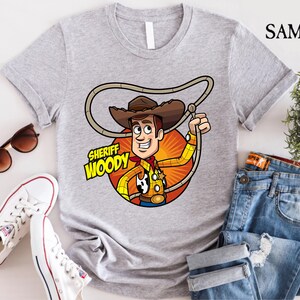 Pode incluir: Uma camiseta cinza com uma imagem de desenho animado do Woody do Toy Story. A imagem mostra o Woody usando seu distintivo de xerife e seu chap&eacute;u. O texto "Sheriff Woody" est&aacute; impresso na camiseta.