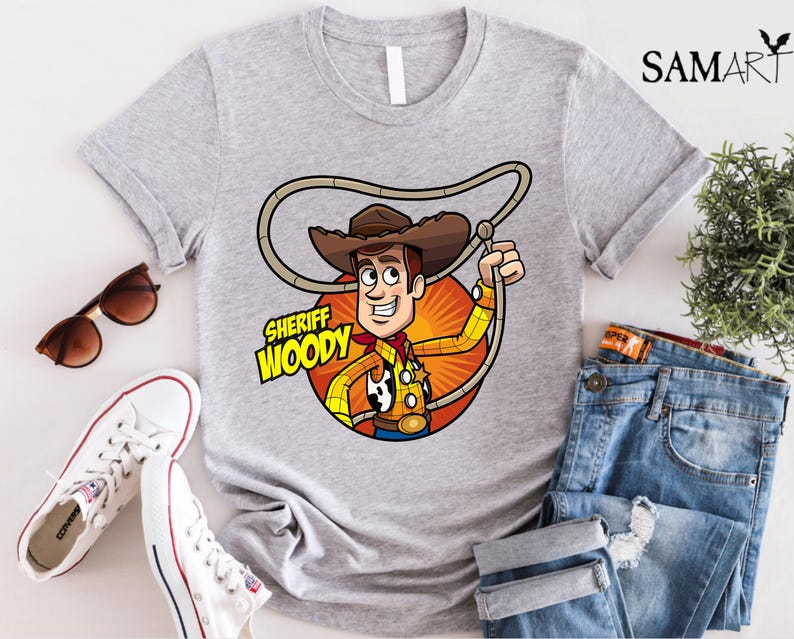 Pode incluir: Uma camiseta cinza com uma imagem de desenho animado do Woody do Toy Story. A imagem mostra o Woody usando seu distintivo de xerife e seu chap&eacute;u. O texto "Sheriff Woody" est&aacute; impresso na camiseta.
