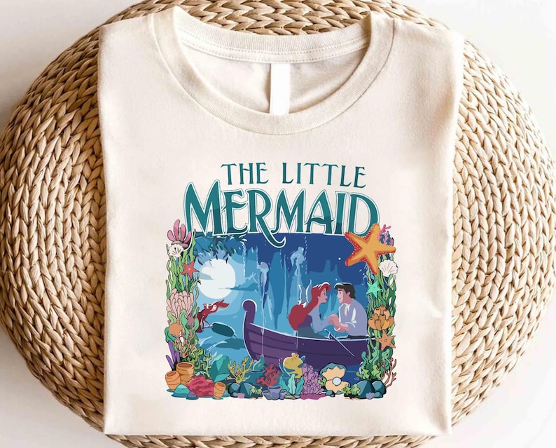 Puede incluir: Camiseta color crema con un gr&aacute;fico colorido de "La Sirenita". El dise&ntilde;o incluye el t&iacute;tulo en verde azulado, un barco con dos personajes y elementos submarinos como coral, una estrella de mar y un cangrejo.