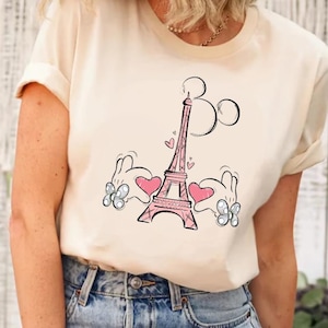 Puede incluir: Una camiseta blanca con un gr&aacute;fico rosa de la Torre Eiffel y manos de Mickey Mouse sosteniendo corazones rosas a cada lado de la torre.
