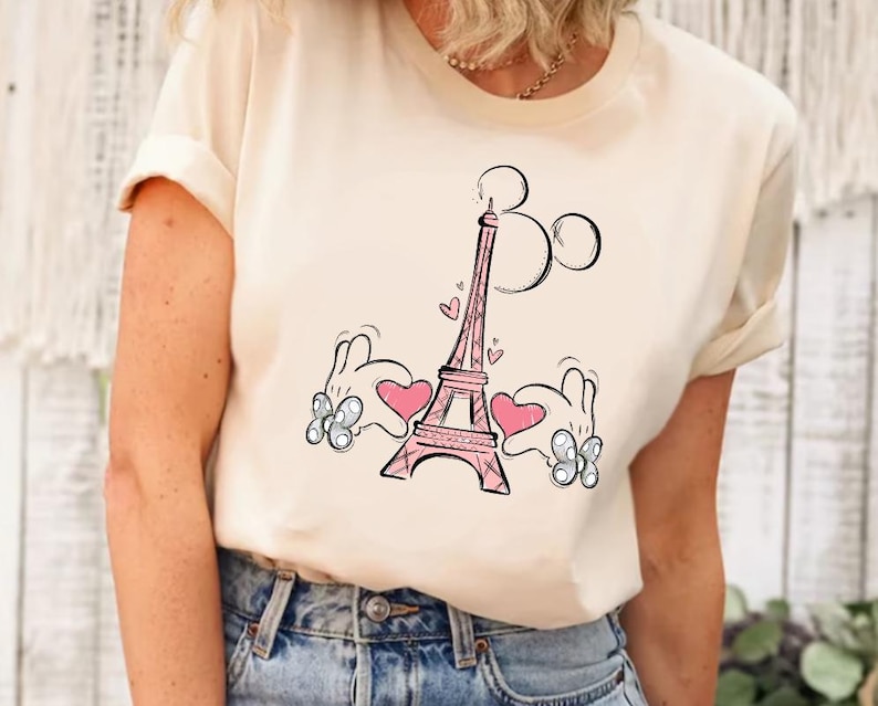 Puede incluir: Una camiseta blanca con un gr&aacute;fico rosa de la Torre Eiffel y manos de Mickey Mouse sosteniendo corazones rosas a cada lado de la torre.