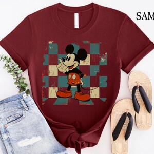 May include: A maroon t-shirt with a vintage Mickey Mouse graphic on a checkered background. The graphic features Mickey Mouse with a red and yellow outfit, giving a thumbs up.