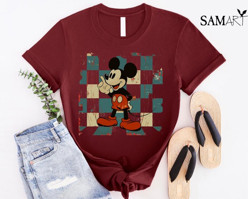 May include: A maroon t-shirt with a vintage Mickey Mouse graphic on a checkered background. The graphic features Mickey Mouse with a red and yellow outfit, giving a thumbs up.