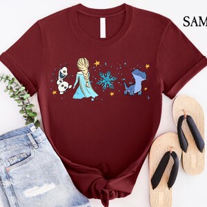 May include: Burgundy t-shirt featuring a graphic of Elsa, Olaf, and a blue dragon with a snowflake and stars. The shirt has a crew neck and short sleeves. The shirt is paired with denim shorts and sandals.