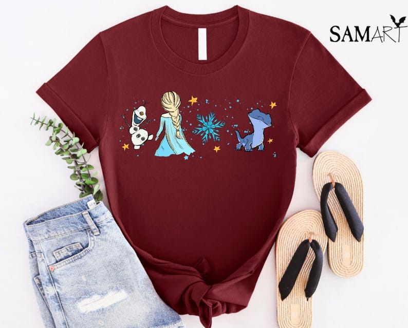 May include: Burgundy t-shirt featuring a graphic of Elsa, Olaf, and a blue dragon with a snowflake and stars. The shirt has a crew neck and short sleeves. The shirt is paired with denim shorts and sandals.