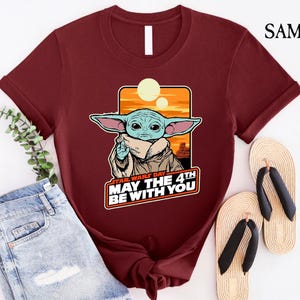 May include: A maroon t-shirt with a cartoon image of a green alien character with big ears. The text "Star Wars Day May the 4th Be With You" is printed on the shirt.