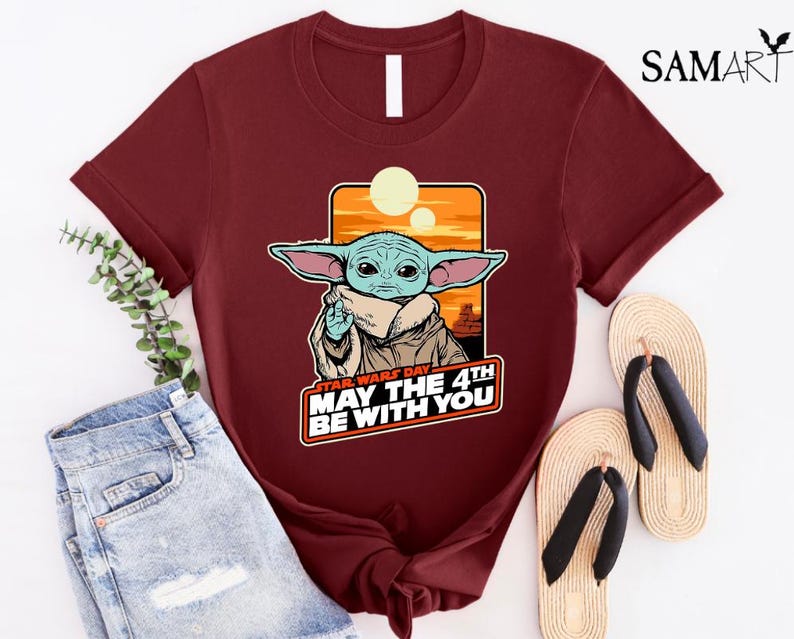May include: A maroon t-shirt with a cartoon image of a green alien character with big ears. The text "Star Wars Day May the 4th Be With You" is printed on the shirt.