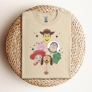 Op de afbeelding: Een beige t-shirt met een afbeelding van gezichten van stripfiguren, waaronder een cowboy, een varken, een ruimteranger, een cowgirl, een hond en een dinosaurus. De karakters zijn gerangschikt als ballonnen. Zwarte sterren accentueren het ontwerp.