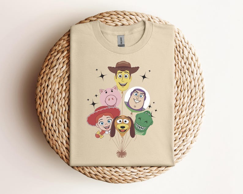 Op de afbeelding: Een beige t-shirt met een afbeelding van gezichten van stripfiguren, waaronder een cowboy, een varken, een ruimteranger, een cowgirl, een hond en een dinosaurus. De karakters zijn gerangschikt als ballonnen. Zwarte sterren accentueren het ontwerp.