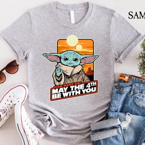 May include: A gray t-shirt with a cartoon image of a green alien character with big ears. The text "Star Wars Day May the 4th Be With You" is printed on the shirt.