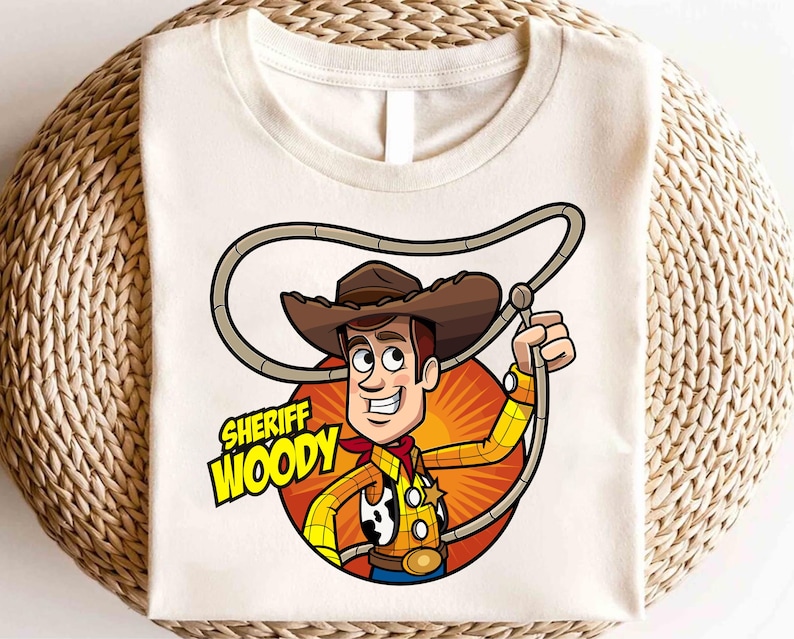 Pode incluir: Uma camiseta branca com uma imagem de desenho animado do Woody do Toy Story. Woody est&aacute; usando seu distintivo de xerife e segurando seu la&ccedil;o. O texto "Sheriff Woody" est&aacute; impresso abaixo da imagem.