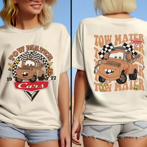 May include: A beige t-shirt with a graphic of Tow Mater from the Cars movie. The graphic features Tow Mater with the text "Tow Mater" and "Cars" in a red circle with a checkered flag design. The text "Tow Mater" is also printed on the back of the shirt.