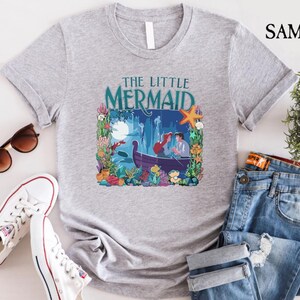 Puede incluir: Una camiseta gris claro con un gr&aacute;fico colorido de "La Sirenita". El dise&ntilde;o incluye a Ariel y el Pr&iacute;ncipe Eric en un bote, rodeados de flora y fauna submarina. Las palabras "THE LITTLE MERMAID" est&aacute;n impresas encima de la imagen en color turquesa.