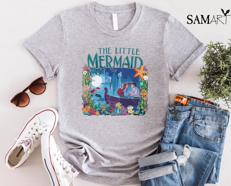 Puede incluir: Una camiseta gris claro con un gr&aacute;fico colorido de "La Sirenita". El dise&ntilde;o incluye a Ariel y el Pr&iacute;ncipe Eric en un bote, rodeados de flora y fauna submarina. Las palabras "THE LITTLE MERMAID" est&aacute;n impresas encima de la imagen en color turquesa.