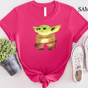 May include: A vibrant pink t-shirt with a graphic of a green alien-like character with large ears. The graphic is set against a desert landscape. White trainers are next to the shirt. The word "SAMART" is in the upper right corner.