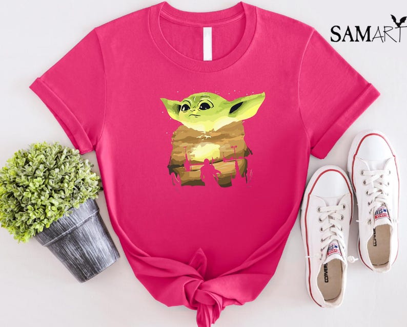 May include: A vibrant pink t-shirt with a graphic of a green alien-like character with large ears. The graphic is set against a desert landscape. White trainers are next to the shirt. The word "SAMART" is in the upper right corner.