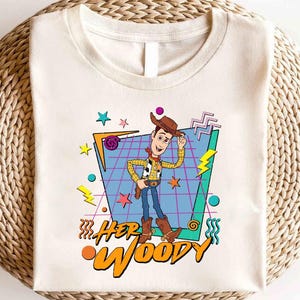 以下が含まれることがあります： トイストーリーのウッディのレトロなグラフィックがプリントされた白いTシャツ。グラフィックは、青いグリッドパターンにカラフルなアクセントを加え、"HER WOODY"というテキストが書かれています。
