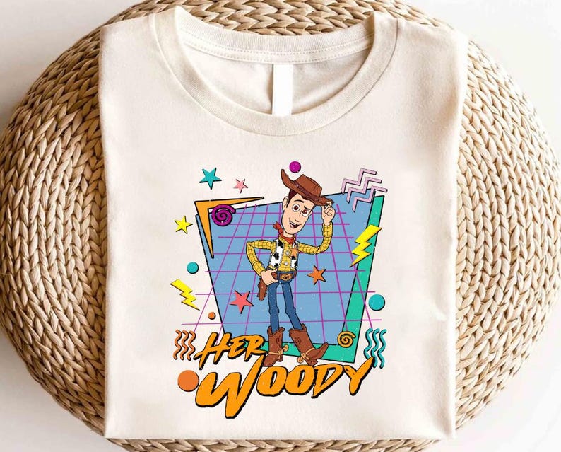 以下が含まれることがあります： トイストーリーのウッディのレトロなグラフィックがプリントされた白いTシャツ。グラフィックは、青いグリッドパターンにカラフルなアクセントを加え、"HER WOODY"というテキストが書かれています。
