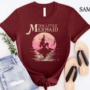 May include: A maroon t-shirt with a vintage-style graphic of Ariel, the Little Mermaid, sitting on a rock with a sunset behind her. The text "The Little Mermaid" is above the graphic.