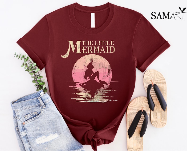 May include: A maroon t-shirt with a vintage-style graphic of Ariel, the Little Mermaid, sitting on a rock with a sunset behind her. The text "The Little Mermaid" is above the graphic.