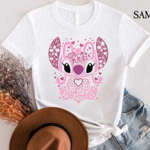 May include: White t-shirt with a pink and white floral design of a cartoon character with big eyes and a heart. The character is surrounded by hearts.