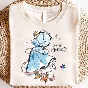 Può includere: Una maglietta color crema con un'immagine di cartone animato di Cenerentola in un abito blu, che tiene un orologio che segna le 'XII'. Il testo 'Dance 'Til Midnight' è stampato sotto l'immagine.