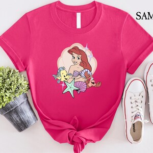 May include: A hot pink t-shirt featuring a cartoon illustration of a mermaid with red hair, a starfish, and two cartoon sea creatures. The shirt is paired with white canvas sneakers. The word "SAMART" is printed in black in the upper right corner.