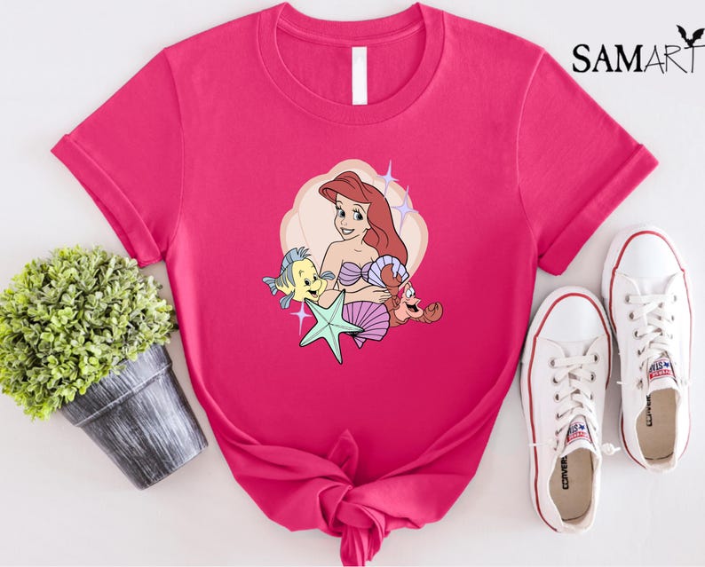 May include: A hot pink t-shirt featuring a cartoon illustration of a mermaid with red hair, a starfish, and two cartoon sea creatures. The shirt is paired with white canvas sneakers. The word "SAMART" is printed in black in the upper right corner.