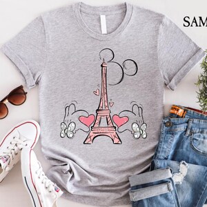 Puede incluir: Una camiseta gris con una ilustraci&oacute;n rosa y negra de la Torre Eiffel y las manos de Mickey Mouse. Las manos sostienen corazones rosas. El texto "Par&iacute;s" est&aacute; escrito sobre la Torre Eiffel.