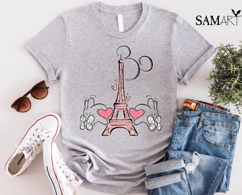 Puede incluir: Una camiseta gris con una ilustraci&oacute;n rosa y negra de la Torre Eiffel y las manos de Mickey Mouse. Las manos sostienen corazones rosas. El texto "Par&iacute;s" est&aacute; escrito sobre la Torre Eiffel.