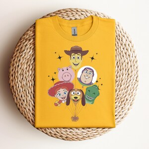 Op de afbeelding: Mosterdgeel T-shirt met gezichten van stripfiguren, waaronder een cowboy, een varken, een ruimteranger, een cowgirl, een hond en een dinosaurus. De karakters zijn gerangschikt als ballonnen. Het shirt is op een geweven, cirkelvormig oppervlak gevouwen.