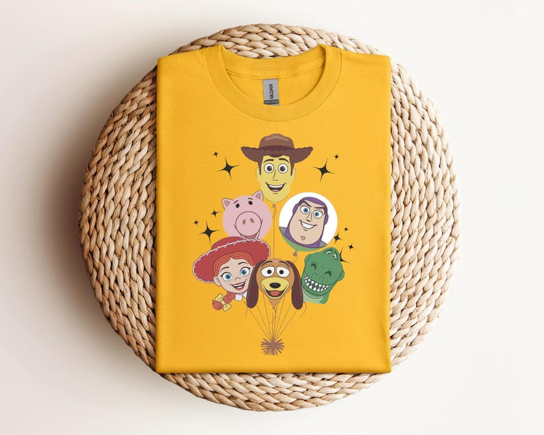 Op de afbeelding: Mosterdgeel T-shirt met gezichten van stripfiguren, waaronder een cowboy, een varken, een ruimteranger, een cowgirl, een hond en een dinosaurus. De karakters zijn gerangschikt als ballonnen. Het shirt is op een geweven, cirkelvormig oppervlak gevouwen.
