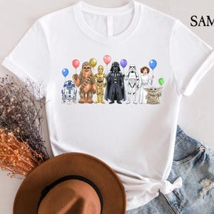 Op de afbeelding: Wit T-shirt met een kleurrijke illustratie van Star Wars-personages die ballonnen vasthouden. De personages zijn R2-D2, Chewbacca, C-3PO, Darth Vader, een Stormtrooper, Princess Leia en Grogu. Op de shirt staat "SAMART" (rechtsboven).