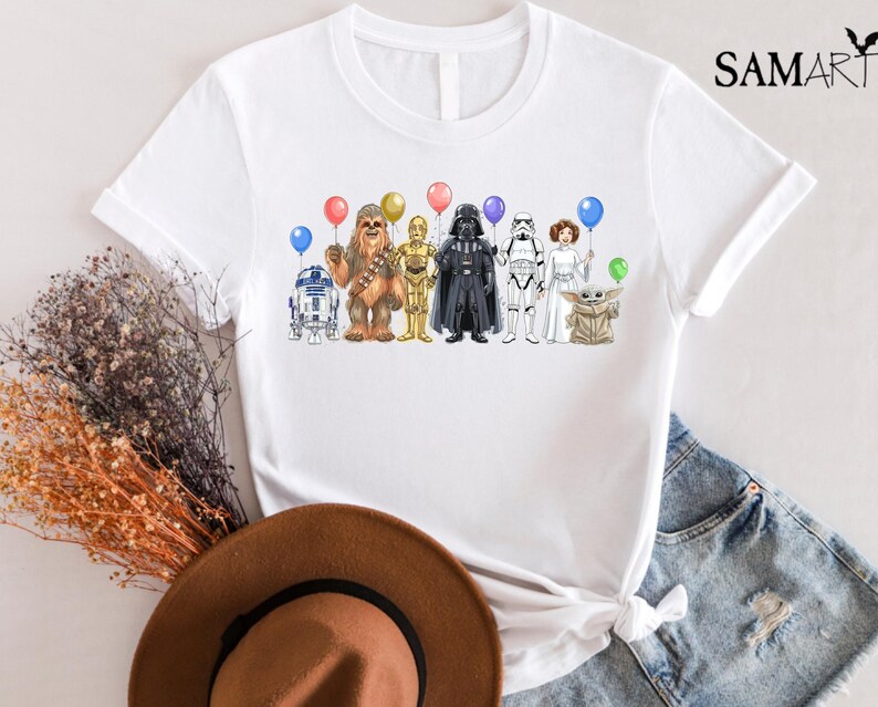 Op de afbeelding: Wit T-shirt met een kleurrijke illustratie van Star Wars-personages die ballonnen vasthouden. De personages zijn R2-D2, Chewbacca, C-3PO, Darth Vader, een Stormtrooper, Princess Leia en Grogu. Op de shirt staat "SAMART" (rechtsboven).