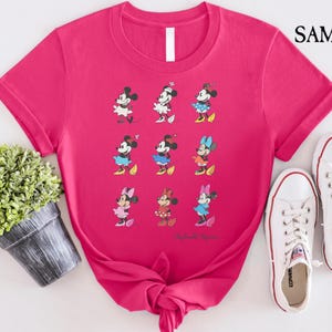 May include: Pink t-shirt with a graphic of nine different Mickey Mouse characters in various poses. The characters are all wearing different colored outfits. The text "Mickey Mouse" is printed at the bottom of the shirt.