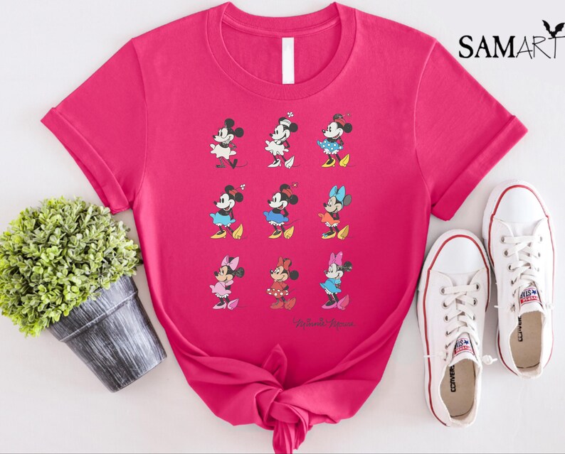 May include: Pink t-shirt with a graphic of nine different Mickey Mouse characters in various poses. The characters are all wearing different colored outfits. The text "Mickey Mouse" is printed at the bottom of the shirt.