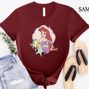 May include: A maroon t-shirt featuring a cartoon illustration of Ariel from The Little Mermaid, with Flounder, Sebastian, and a starfish. The design is set against a cream-colored cloud shape. The shirt is paired with denim shorts and sandals.
