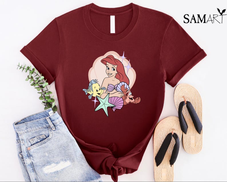 May include: A maroon t-shirt featuring a cartoon illustration of Ariel from The Little Mermaid, with Flounder, Sebastian, and a starfish. The design is set against a cream-colored cloud shape. The shirt is paired with denim shorts and sandals.