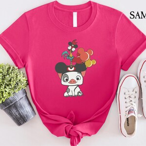 May include: A hot pink t-shirt featuring a cartoon illustration of a pig with a rooster and balloons. The shirt has a crew neck and short sleeves. White sneakers are next to the shirt. The text "SAMART" is in the upper right corner.