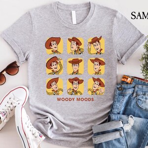 以下が含まれることがあります： トイストーリーのウッディのグラフィックがプリントされたグレーのTシャツ。グラフィックはウッディの顔を9つの異なる表情で示しており、その下に「ウッディムード」というテキストが書かれています。