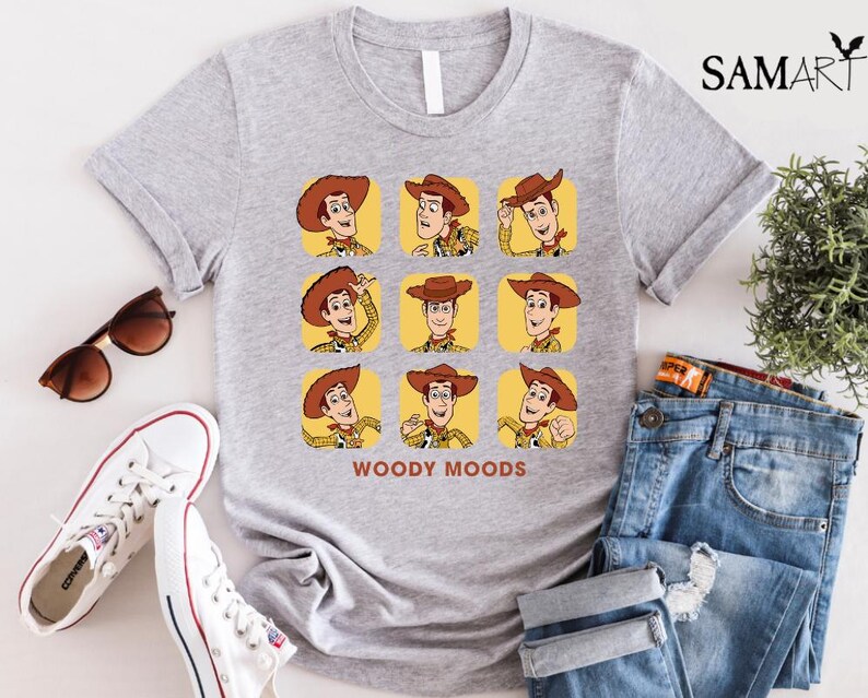 以下が含まれることがあります： トイストーリーのウッディのグラフィックがプリントされたグレーのTシャツ。グラフィックはウッディの顔を9つの異なる表情で示しており、その下に「ウッディムード」というテキストが書かれています。