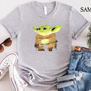 May include: A light grey t-shirt with a graphic of a green alien character with large ears. The character is set against a brown and yellow background. The shirt is paired with blue jeans, white trainers, and sunglasses.