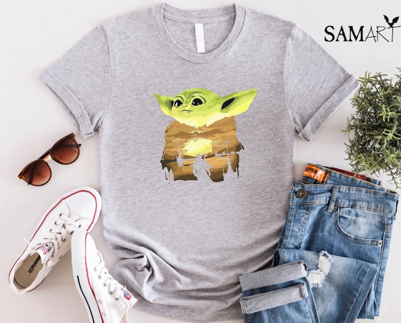 May include: A light grey t-shirt with a graphic of a green alien character with large ears. The character is set against a brown and yellow background. The shirt is paired with blue jeans, white trainers, and sunglasses.