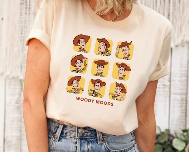 以下が含まれることがあります： ウッディの表情が異なるグラフィックがプリントされた白いTシャツ。グラフィックの下に「WOODY MOODS」というテキストがプリントされています。